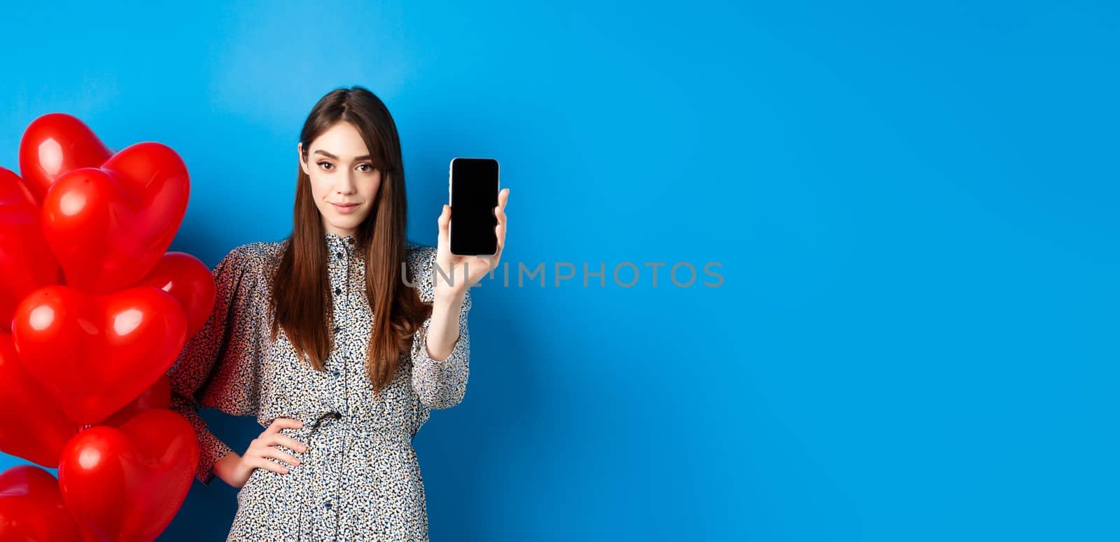 Valentines day. Beautiful young woman with makeup, wearing trendy dress, standing near red hearts balloons and showing empty mobile screen, blue background by Benzoix