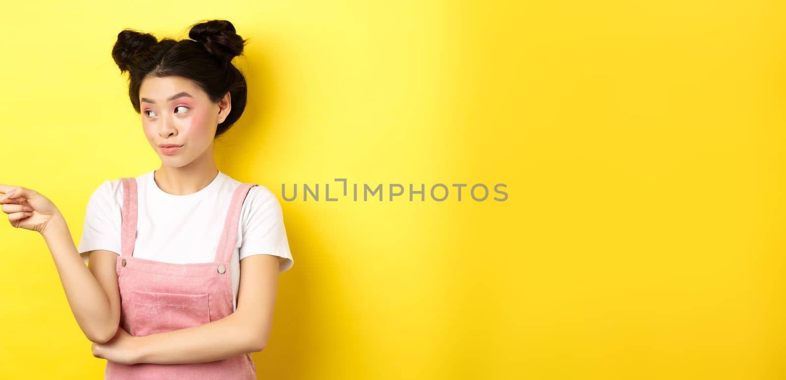 Portrait of stylish asian woman with makeup and summer clothes, pointing and looking left at logo, standing intrigued on yellow background.