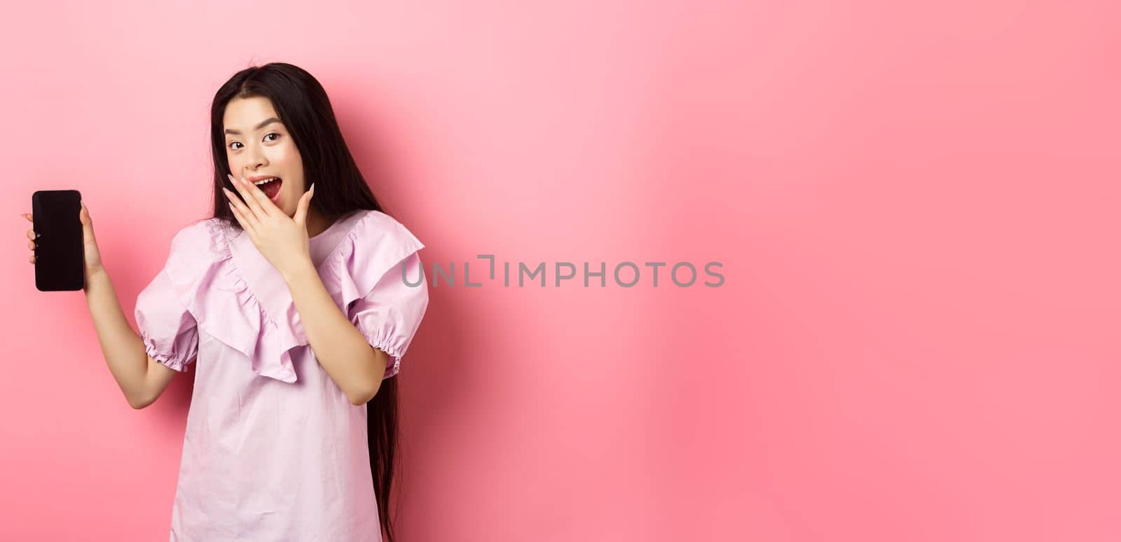 Cheerful asian teen girl showing empty phone screen, laughing and covering mouth with hand, standing in dress against pink background by Benzoix