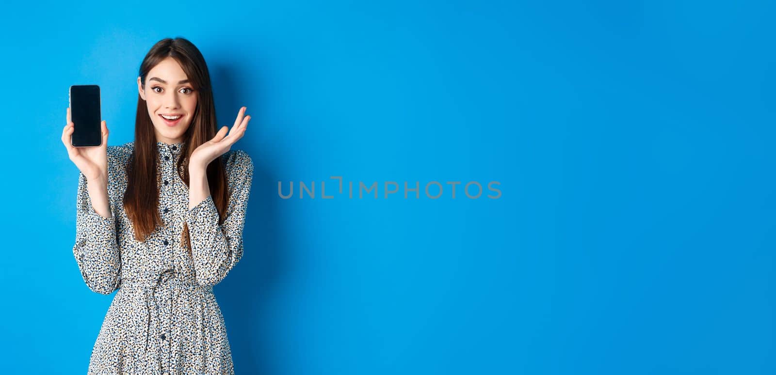 Excited girl showing empty smartphone screen and gasping fascinated, demonstrate shopping app, standing on blue background.