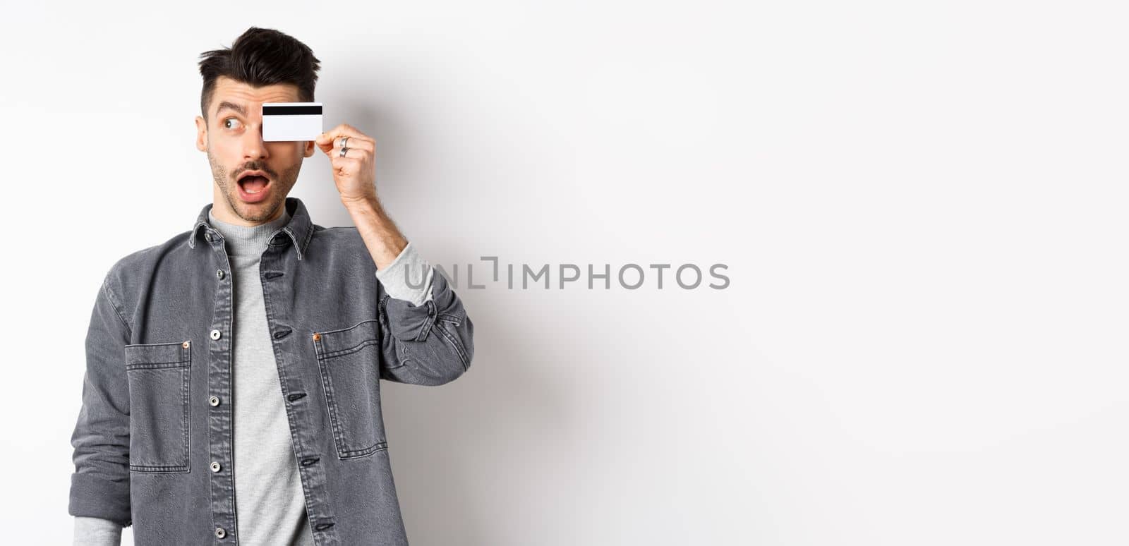 Excited funny guy look aside at logo and show plastic credit card, standing amazed on white background.