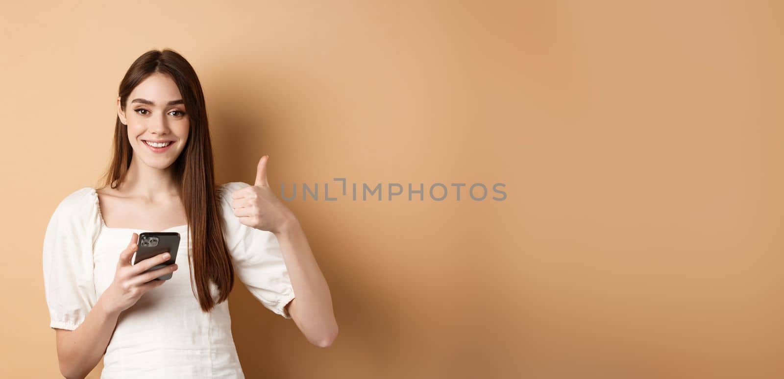 Online shopping concept. Smiling happy woman showing thumb up after using smartphone, holding phone and say yes, standing on beige background.