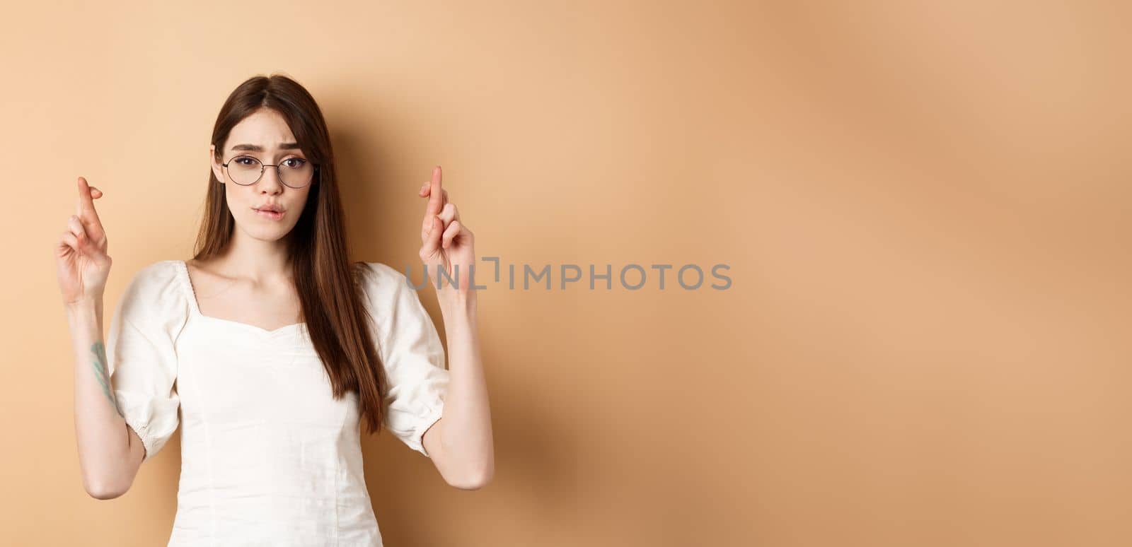 Hopeful girl in glasses cross fingers for good luck, biting lip nervous and looking at camera, waiting for results, praying or making wish, beige background by Benzoix