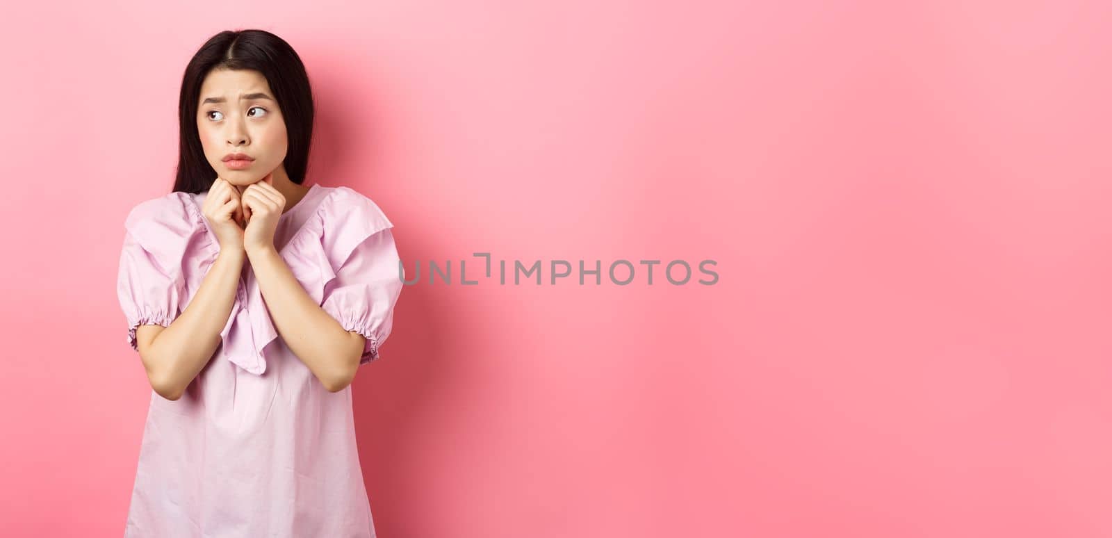 Worried timid asian girl looking aside, holding hands pressed to chest and look concerned, standing sad and gloomy on white background.