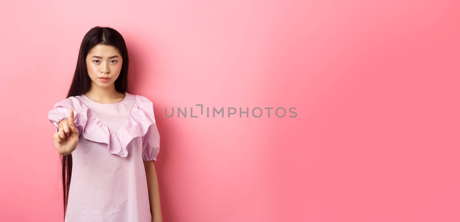 She says no. Serious asian girl shaking finger in stop gesture, prohibit and disagree with person, scolding bad behaviour, standing against pink background by Benzoix