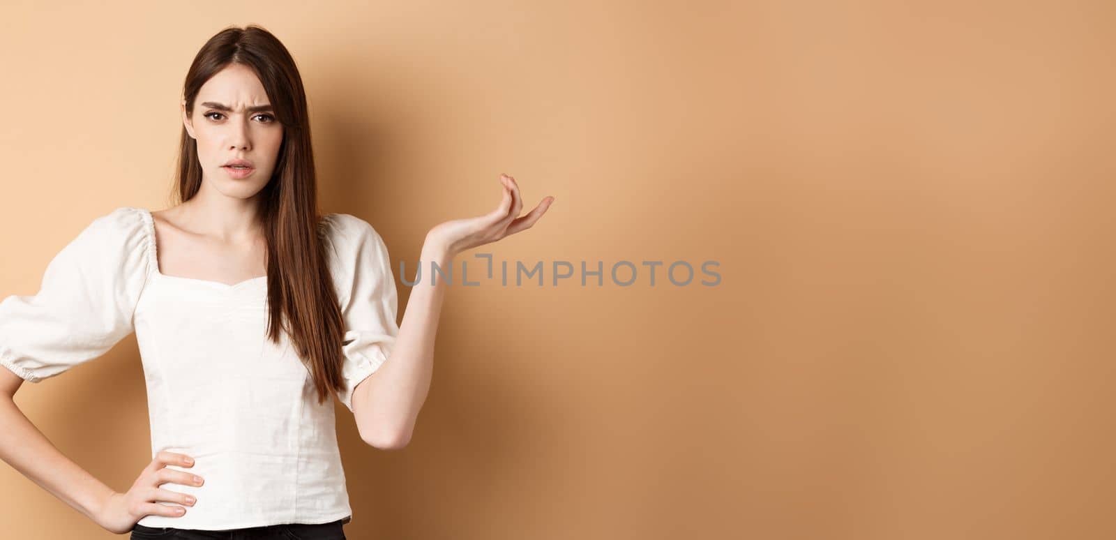 So what. Confused and annoyed young woman raising hand up and staring bothered at camera, cant understand wtf going on, standing on beige background by Benzoix
