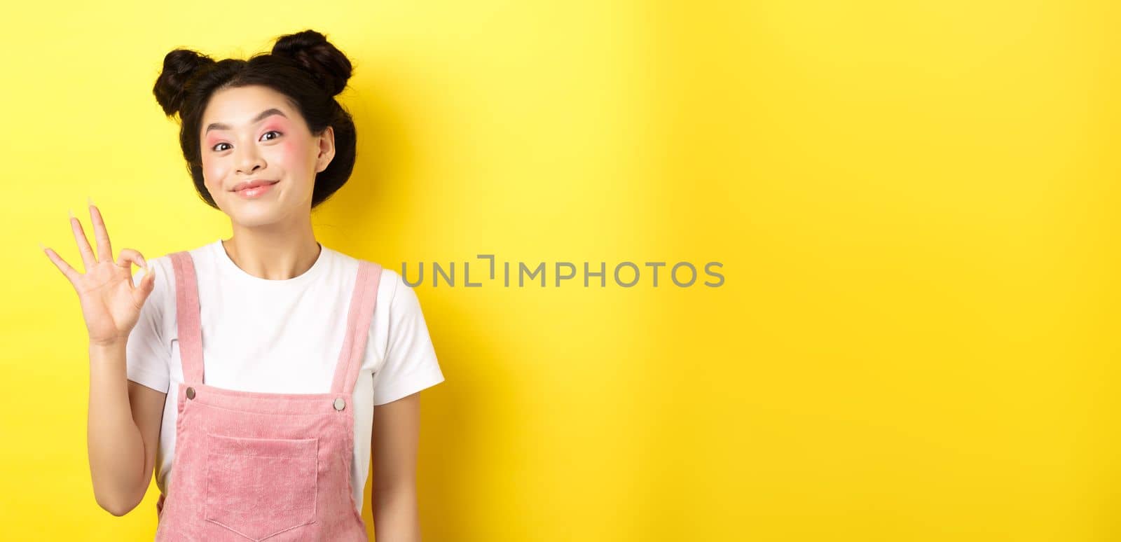Cheerful asian beauty girl with makeup, show okay sign and smiling excited, approve good promo, recommending product, yellow background.