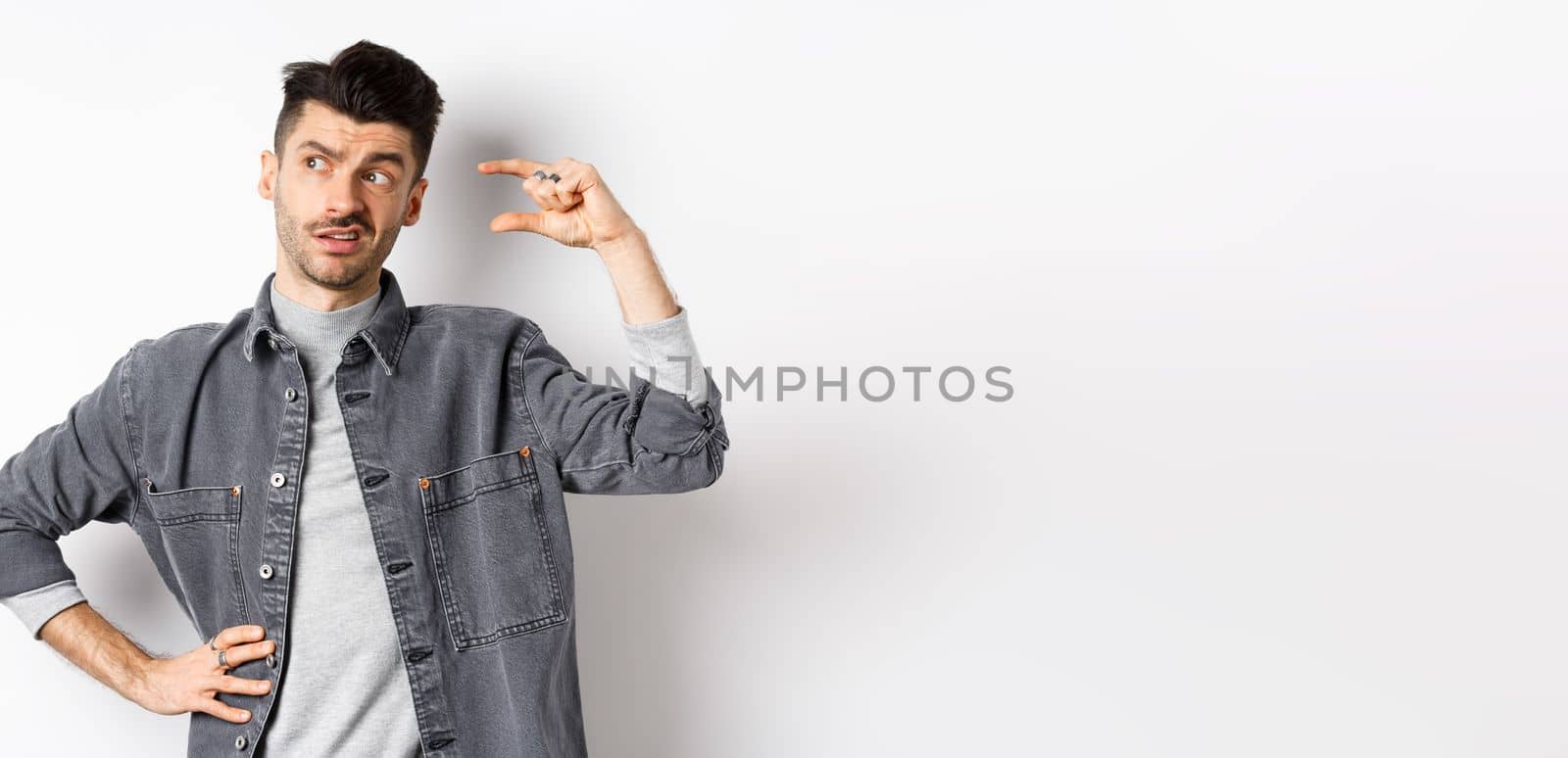 Pensive young man looking aside and thinking, showing small size object, pondering something little or tiny, standing on white background by Benzoix