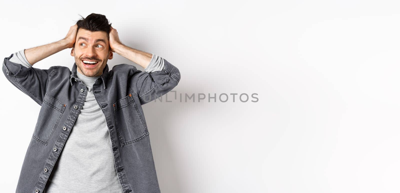 Excited young man looking relieved and happy aside, smiling and staring at logo, holding hands on head cheerful, standing on white background by Benzoix