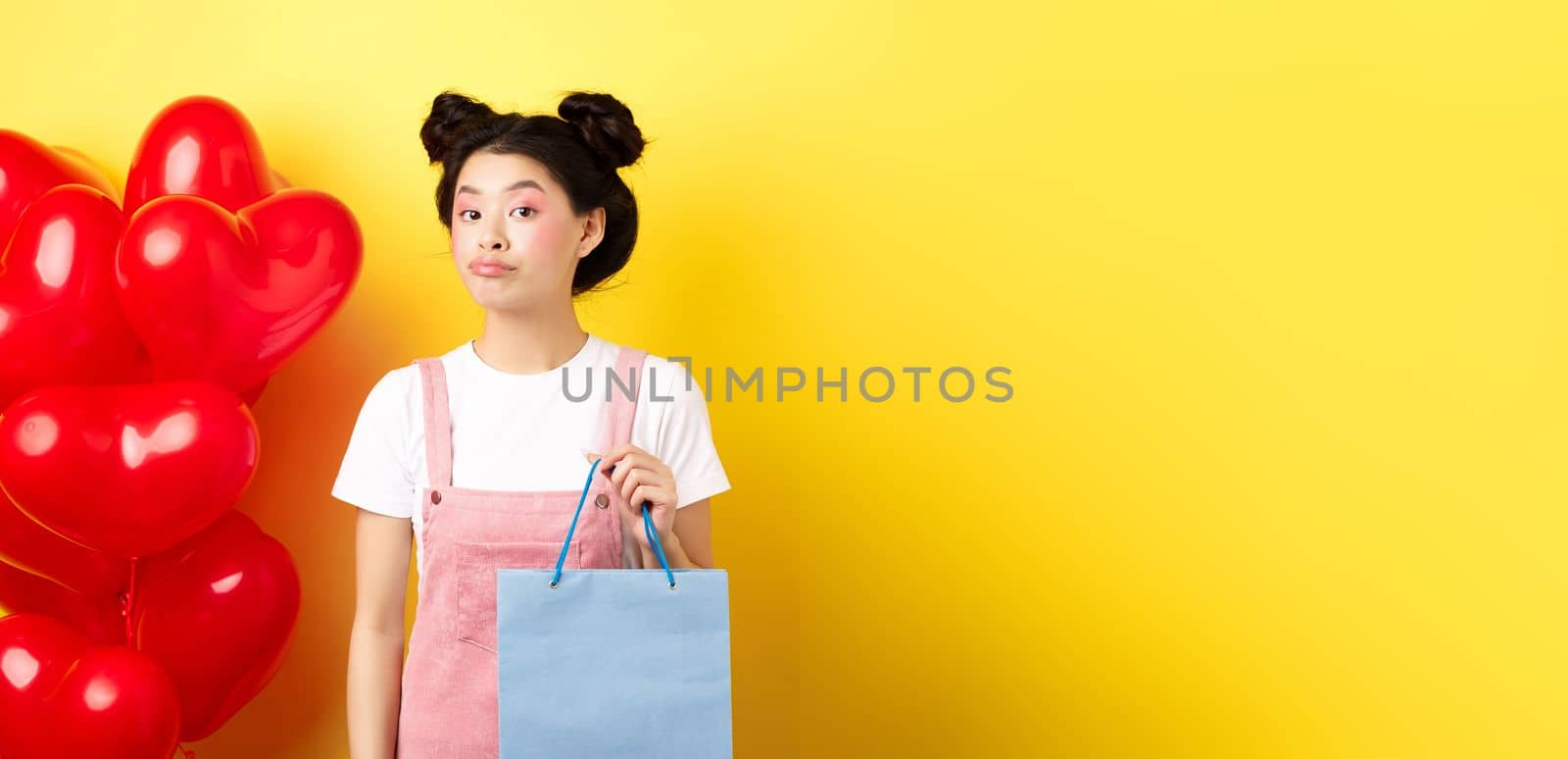 Happy Valentines day. Stylish asian single girl buying herself gift, holding shopping bag and looking unbothered at camera, standing over yellow background.