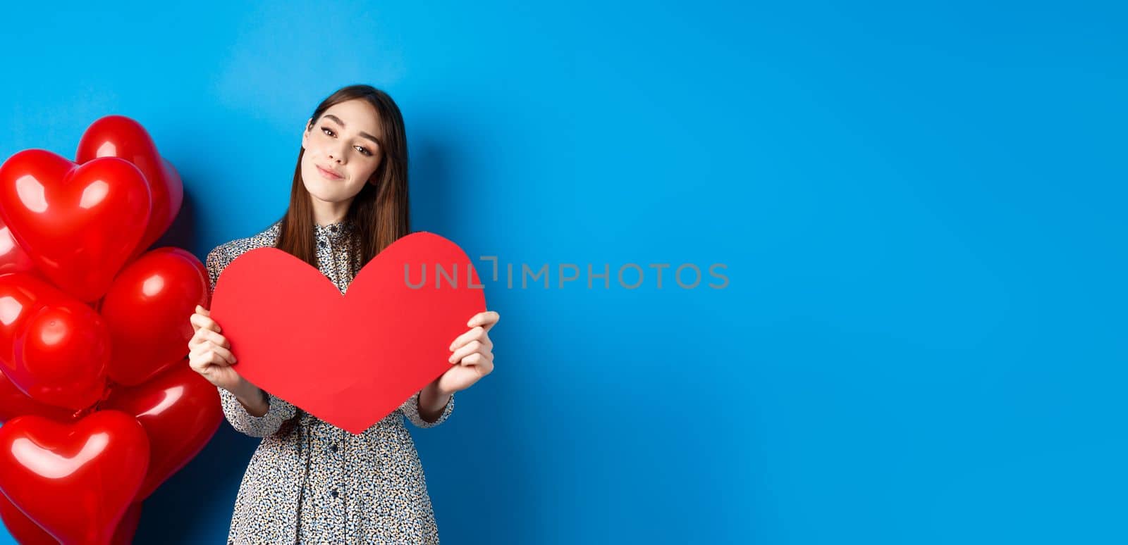 Valentines day. Romantic girl in dress showing big red heart cutout, dreaming of love, standing near holiday balloons on blue background.