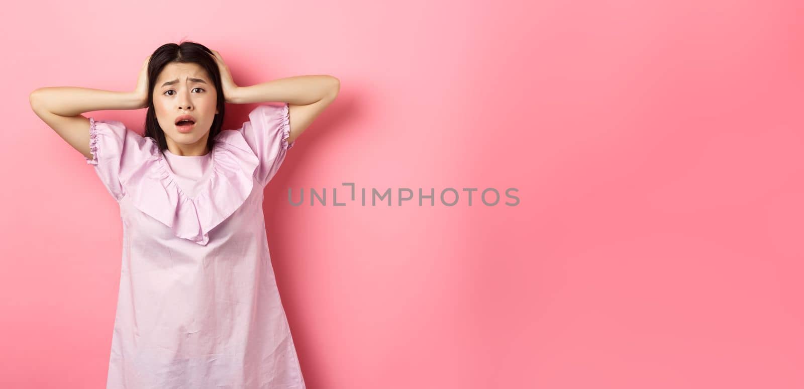 Frustrated asian teen girl panicking, holding hands on head and screaming scared, standing anxious on pink background by Benzoix