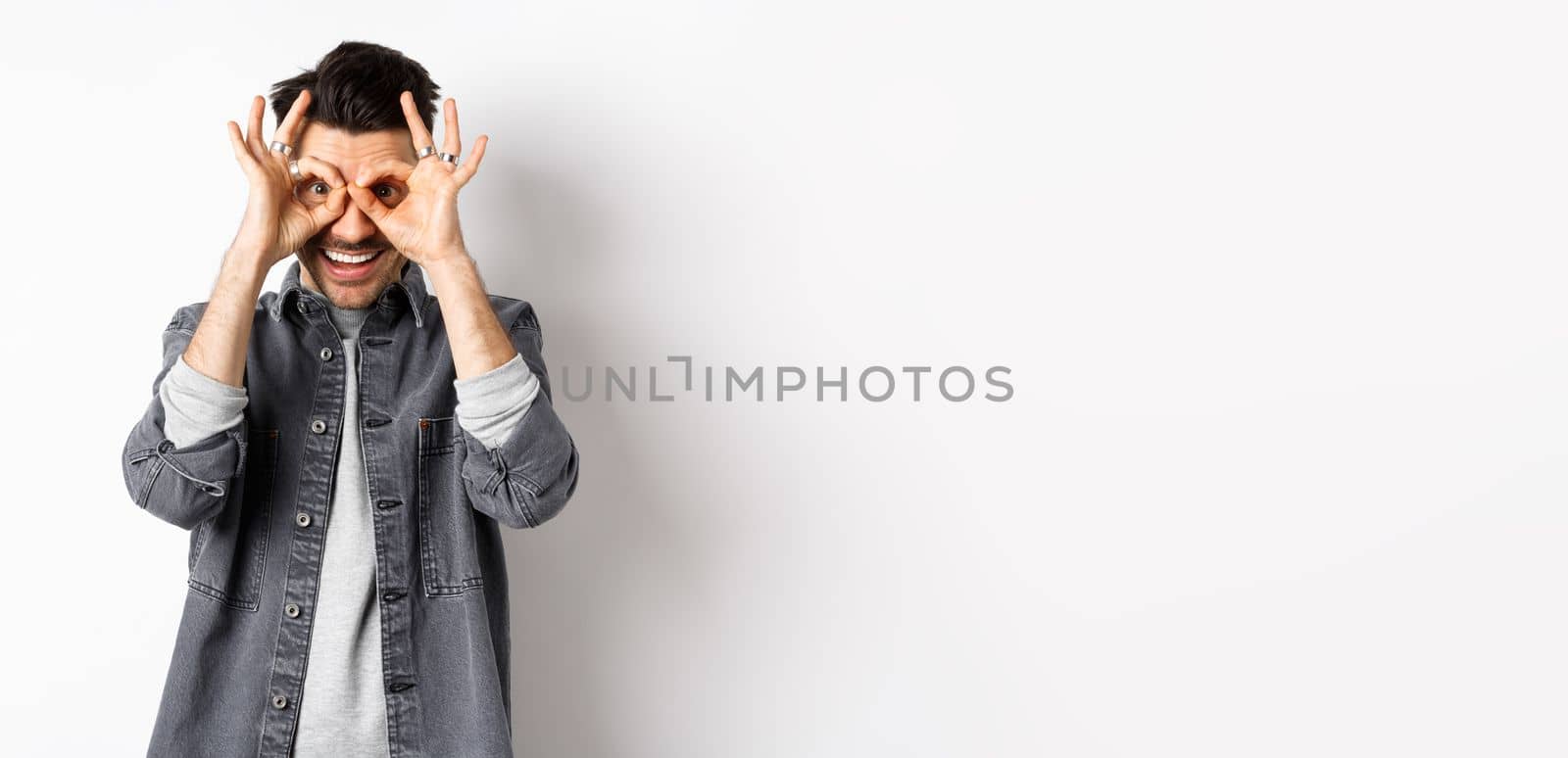 Funny smiling man seeing good offer through hand binoculars, looking happy and excited, checking out advertisement, standing on white background.