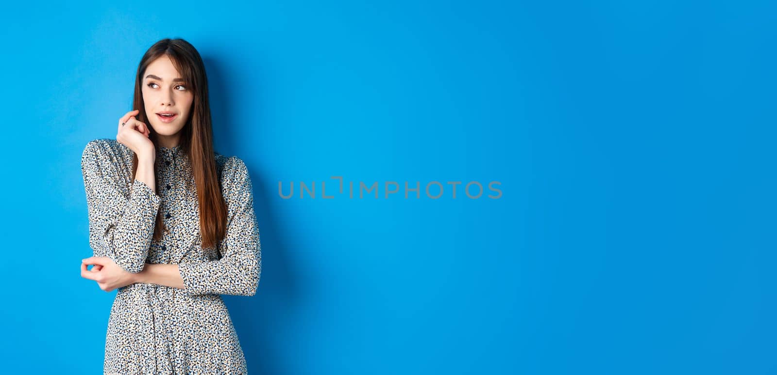 Pensive caucasian girl with long natural hair, wearing vintage dress, looking left with thoughtful face, standing on blue background.