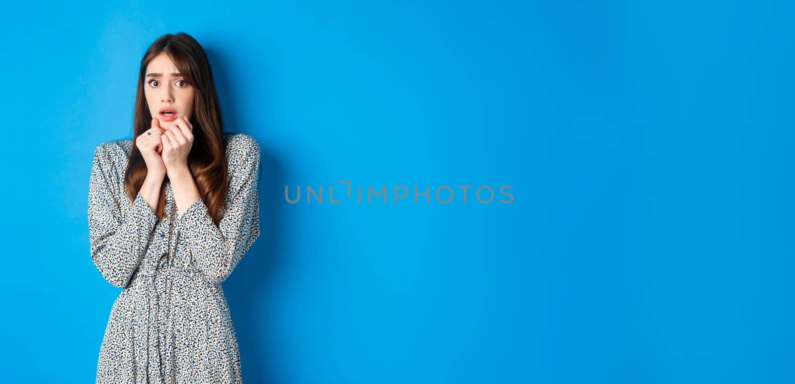 Scared and timid woman in dress, shaking from fear, gasping with hands pressed to chest, looking at scary horror movie, standing against blue background by Benzoix