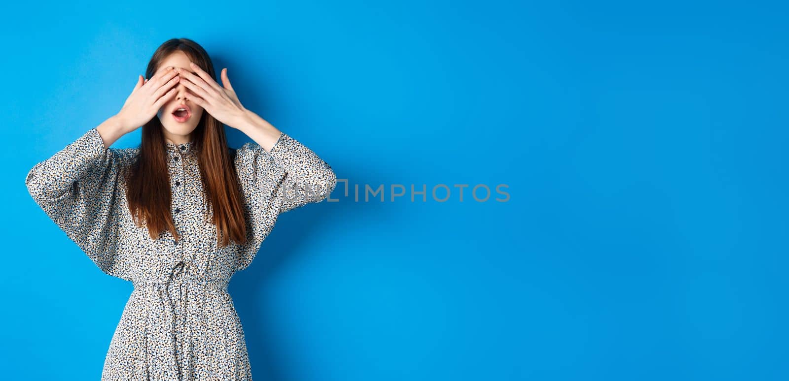 Excited girl waiting for surprise while eyes covered with hands, standing intrigued on blue background, wearing dress.
