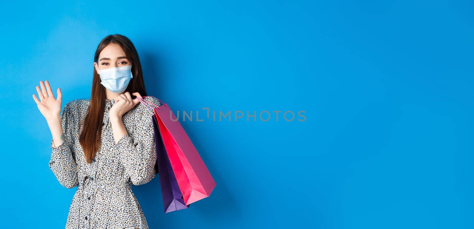 Covid-19, pandemic and lifestyle concept. Friendly cute woman wear medical mask in shopping mall, waving hand and holding bags, say hello, standing on blue background.