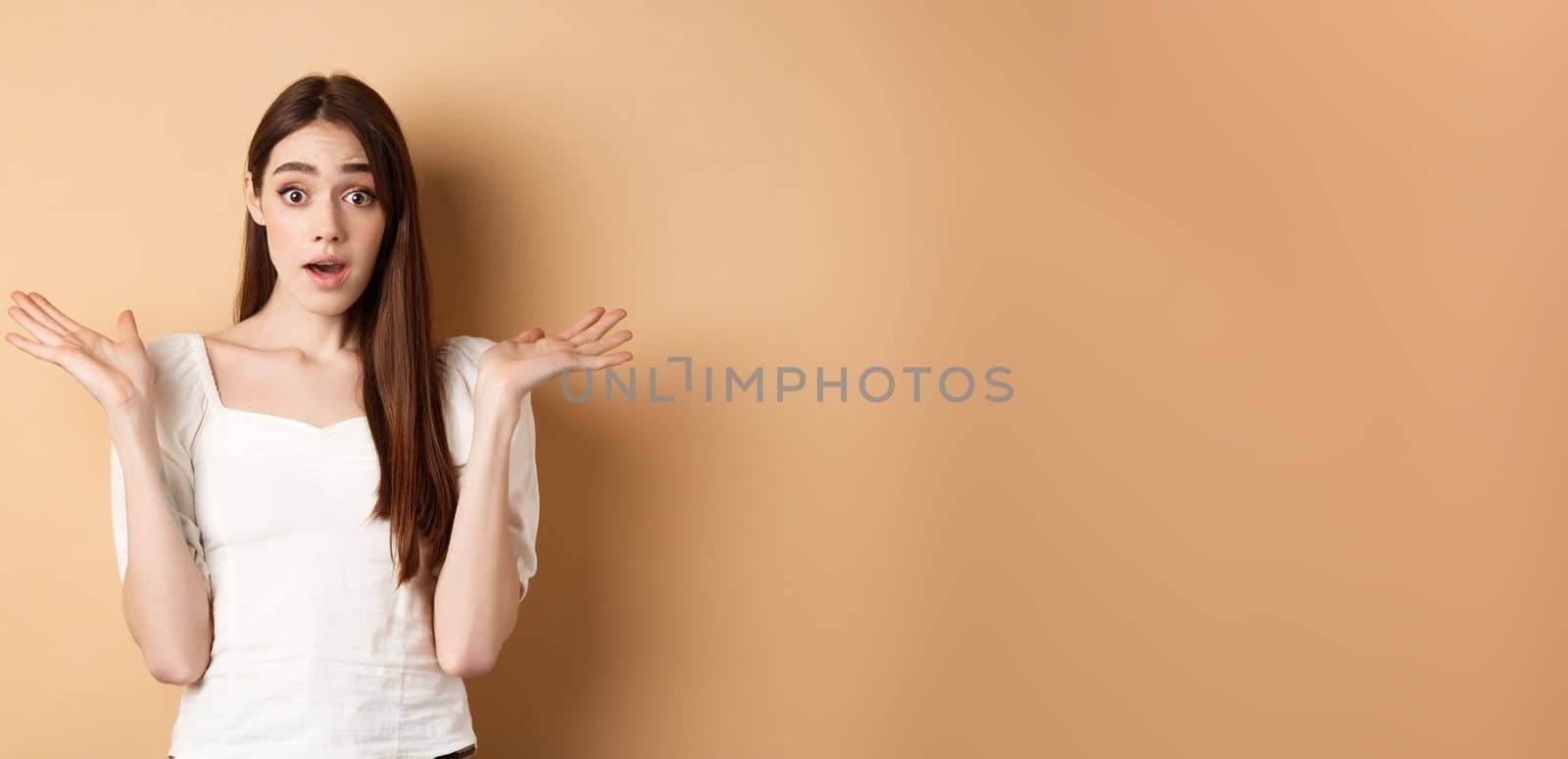 Surprised caucasian girl look with disbelief and amazement, spread hands sideways and gasping confused, cant understand what happening, know nothing, standing on beige background by Benzoix