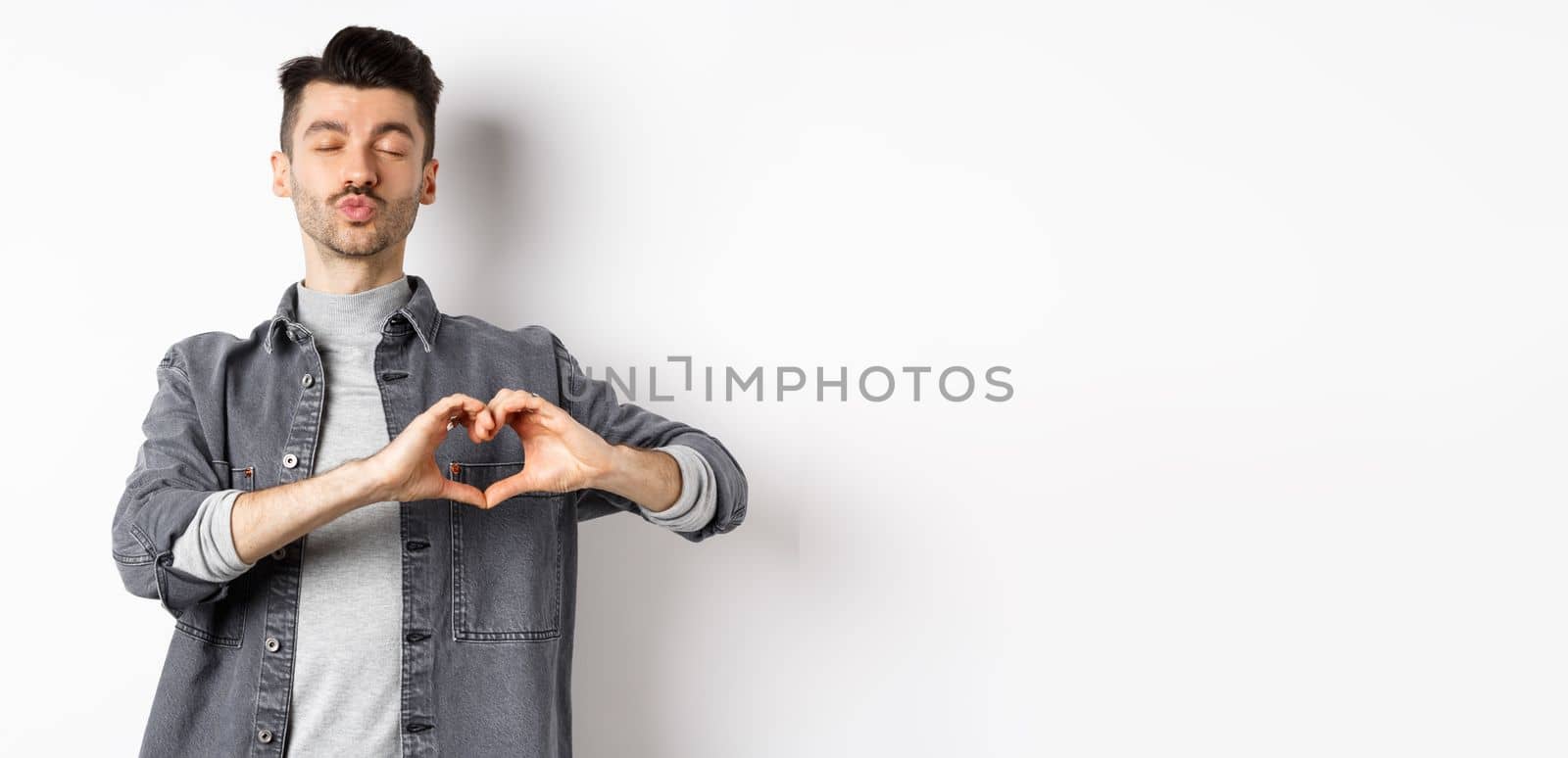 Cute young man waiting for kiss and showing heart gesture, confess on Valentines day, I love you sign, standing against white background.