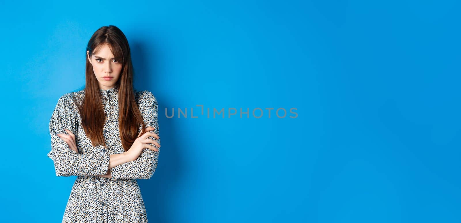 Angry young woman in dress feel unfair, cross arms on chest and frowning offended, standing on blue background by Benzoix