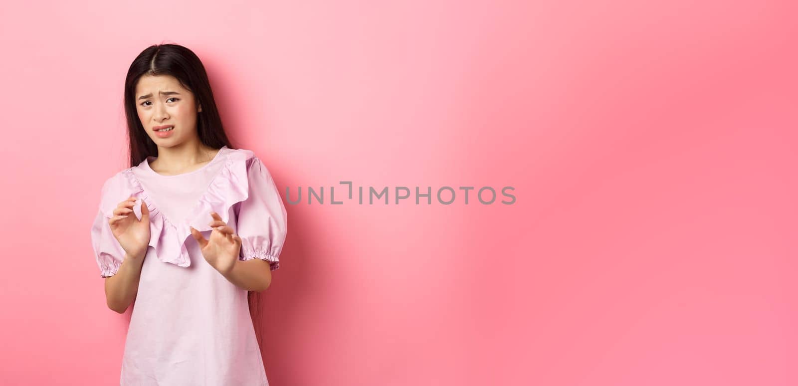Stay away from me. Disgusted asian girl raising hands to block someone, cringe from aversion, look reluctant and asking to stop, rejecting something bad, pink background.