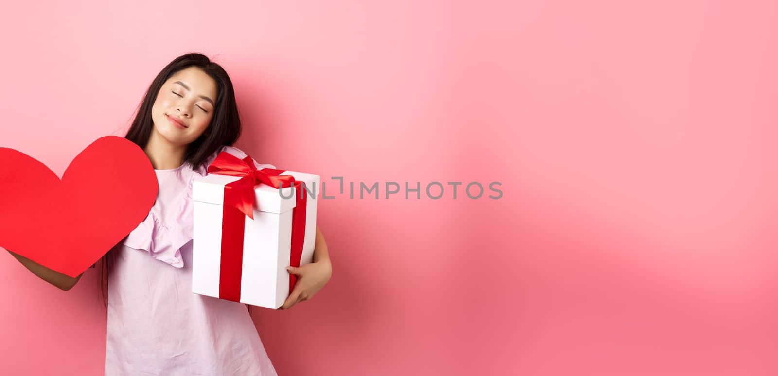 Valentines day concept. Romantic and tender asian teen girl smiling, close eyes and standing dreamy with lover gifts, holding present and big red heart cutout, pink background.