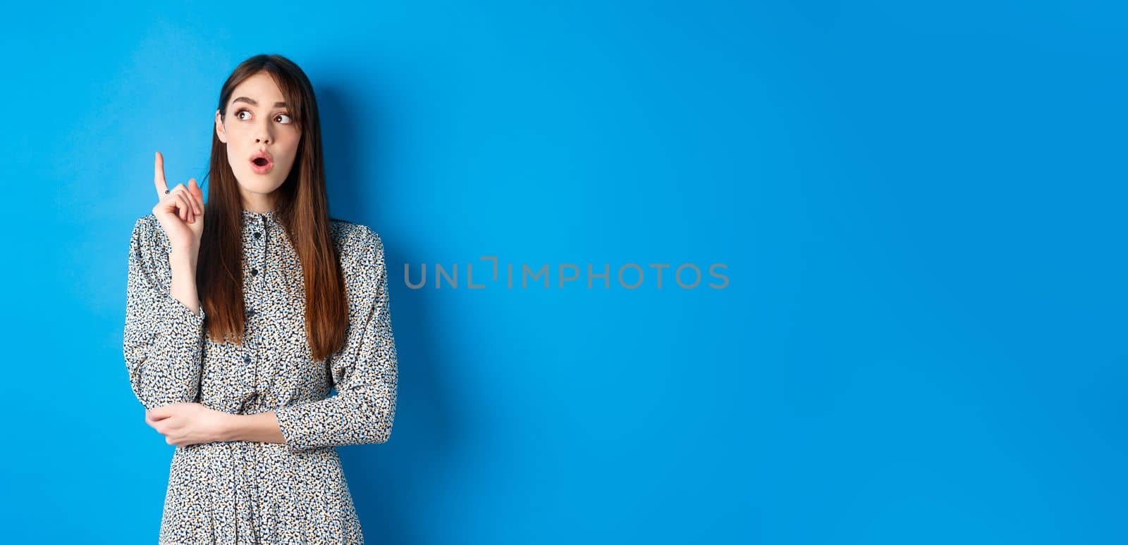 Thoughtful attractive girl in dress pitching an idea, raising finger and looking up, have a plan, standing on blue background by Benzoix