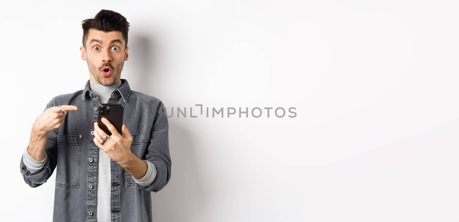 Wow check it out. Excited handsome man showing online offer on phone, pointing at smartphone and looking amazed, standing on white background by Benzoix