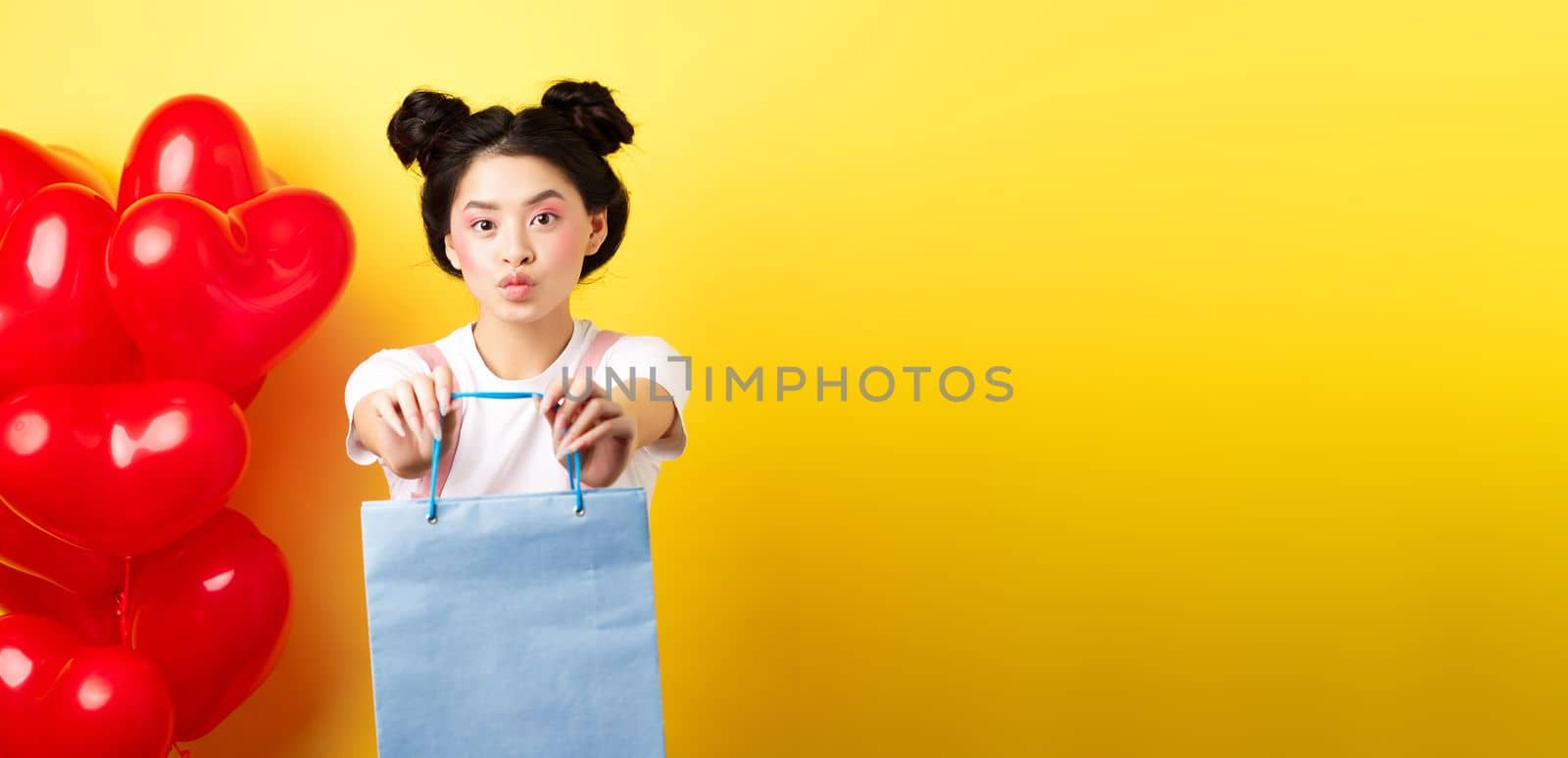 Happy Valentines day. Cute asian girlfriend giving lover a gift, stretch out hands with shopping bag and looking silly, standing over yellow background near heart balloons.