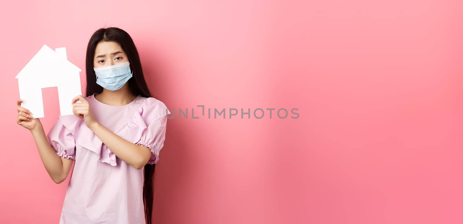 Real estate and pandemic concept. Gloomy girl showing paper house cutout, wearing medical mask and dress, standing against pink background by Benzoix