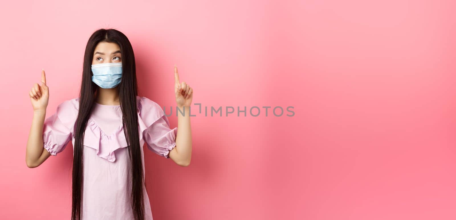 Coronavirus, quarantine and lifestyle concept. Cute korean girl in medical mask pointing fingers up, looking at top advertisement, standing against pink background.