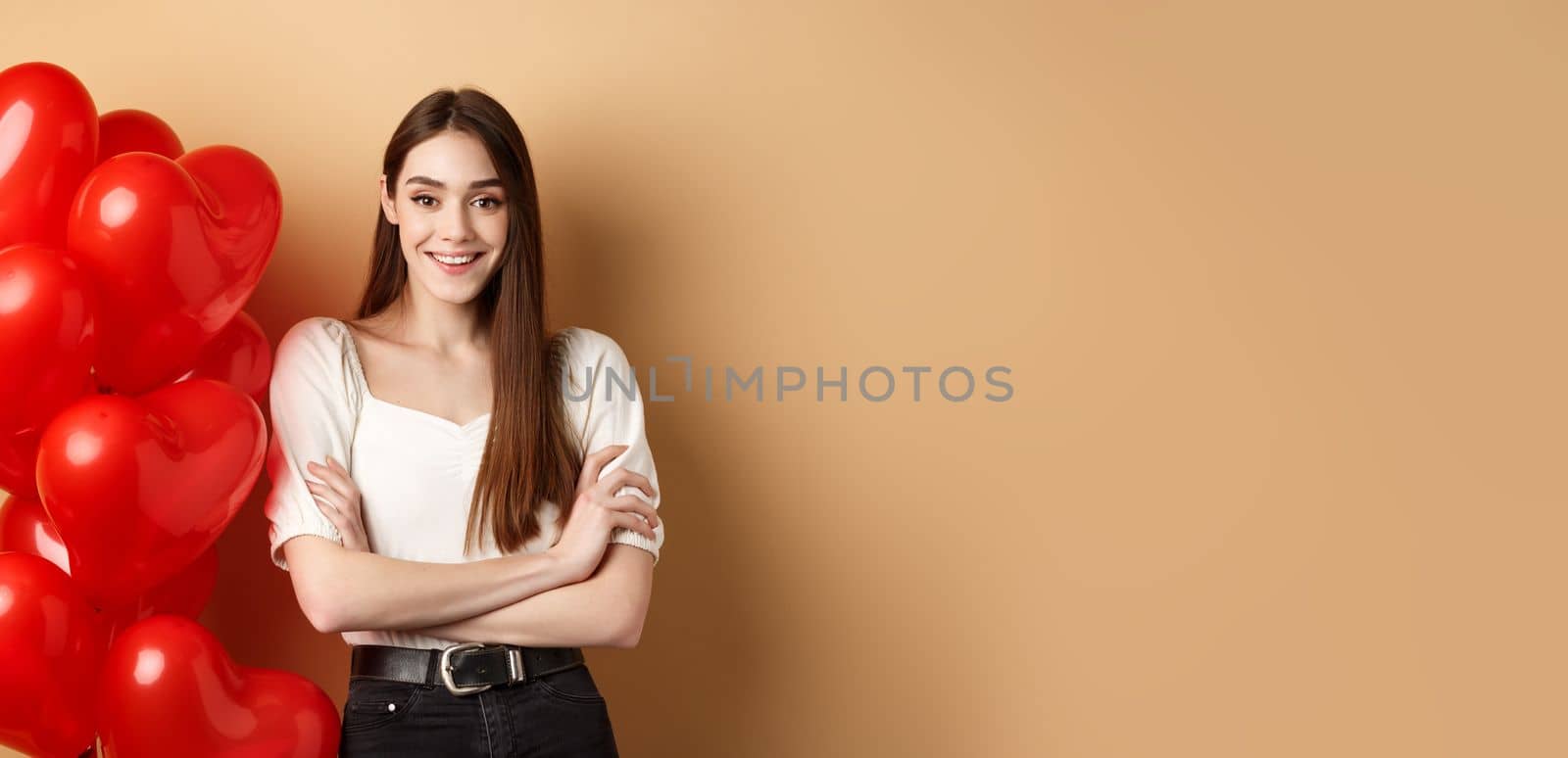 Cheerful young woman looking happy on Valentines day, standing near hearts balloons with arms crossed, smiling at camera, standing on beige background.