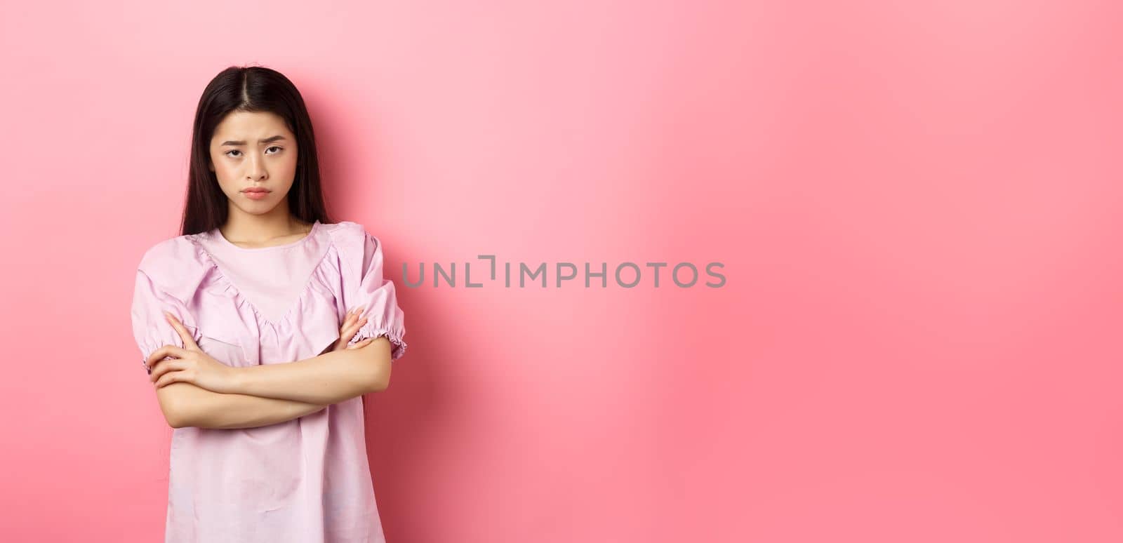Silly asian girl feel unfair, cross arms on chest and sulking, frowning at camera, standing offended against pink background by Benzoix