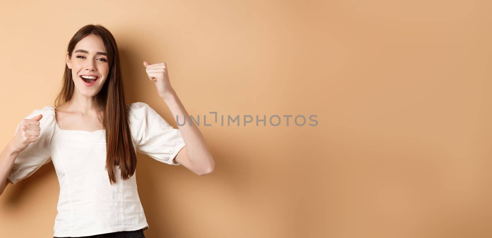 Girl getting motivation, making fist pumps and chanting yes, smiling happy, celebrating victory or cheering, standing on beige background.