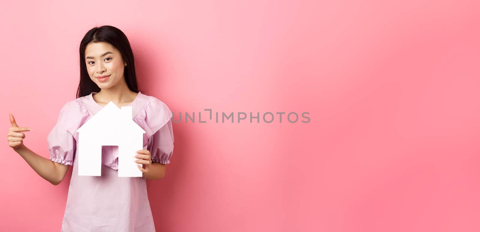 Real estate and insurance concept. Beautiful asian woman pointing at paper house cutout, showing agency logo, standing in dress on pink background by Benzoix