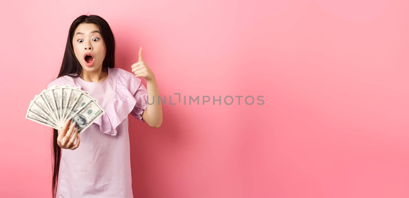 Excited teen girl holding big amount of money, showing dollar bills and thumbs up, standing amazed on pink background by Benzoix