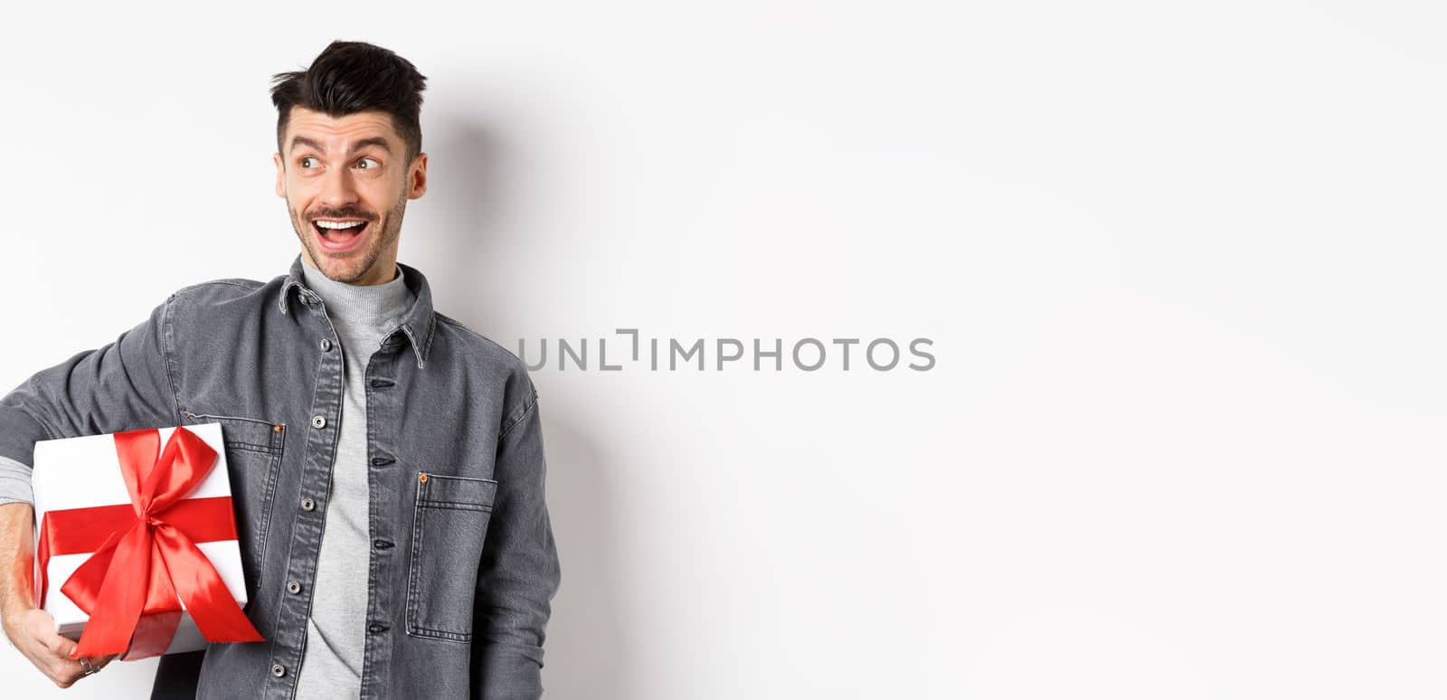 Cheerful handsome guy looking at empty space with amazed smile, checking out special valentines day offer, holding big romantic gift box, standing on white background.