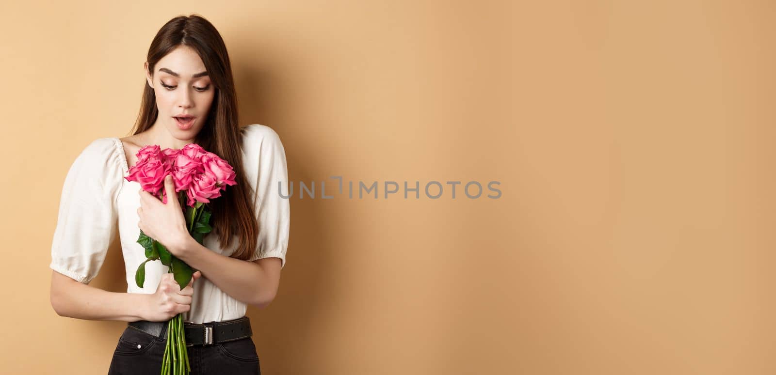 Valentines day. Surprised girl receive gift from lover on date, looking amazed at beautiful bouquet of flowers, holding roses on beige background.