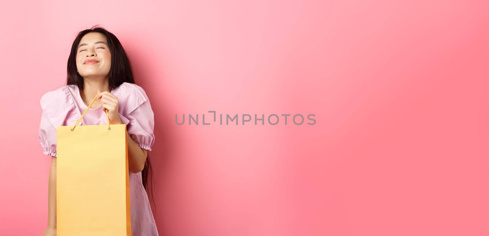 Beautiful happy asian woman holding shopping bag, smiling with eyes closed, standing in dress against pink background.