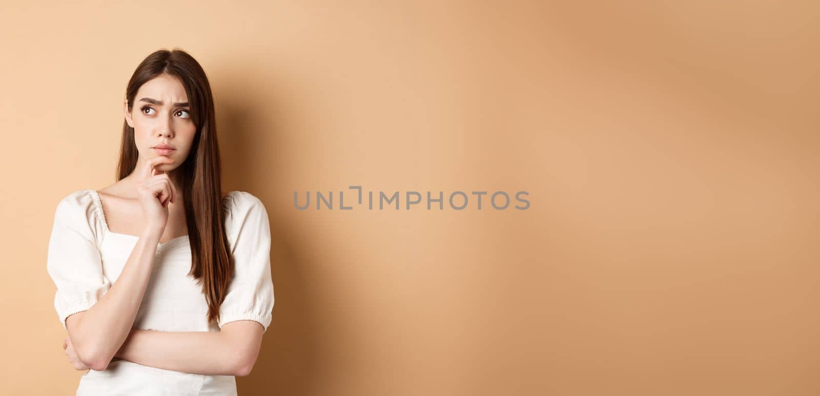 Worried young woman staring pensive aside, touching chin and thinking, pondering plan, looking for ideas, standing on beige background by Benzoix