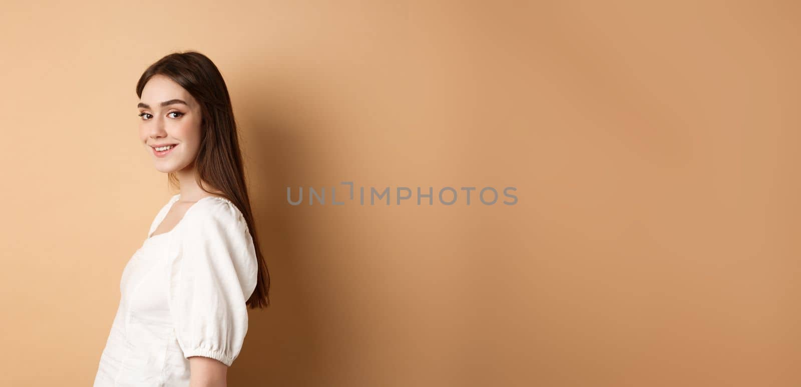 Profile of pretty european girl turn head at camera and smiling coquettish, standing in white blouse against beige background by Benzoix