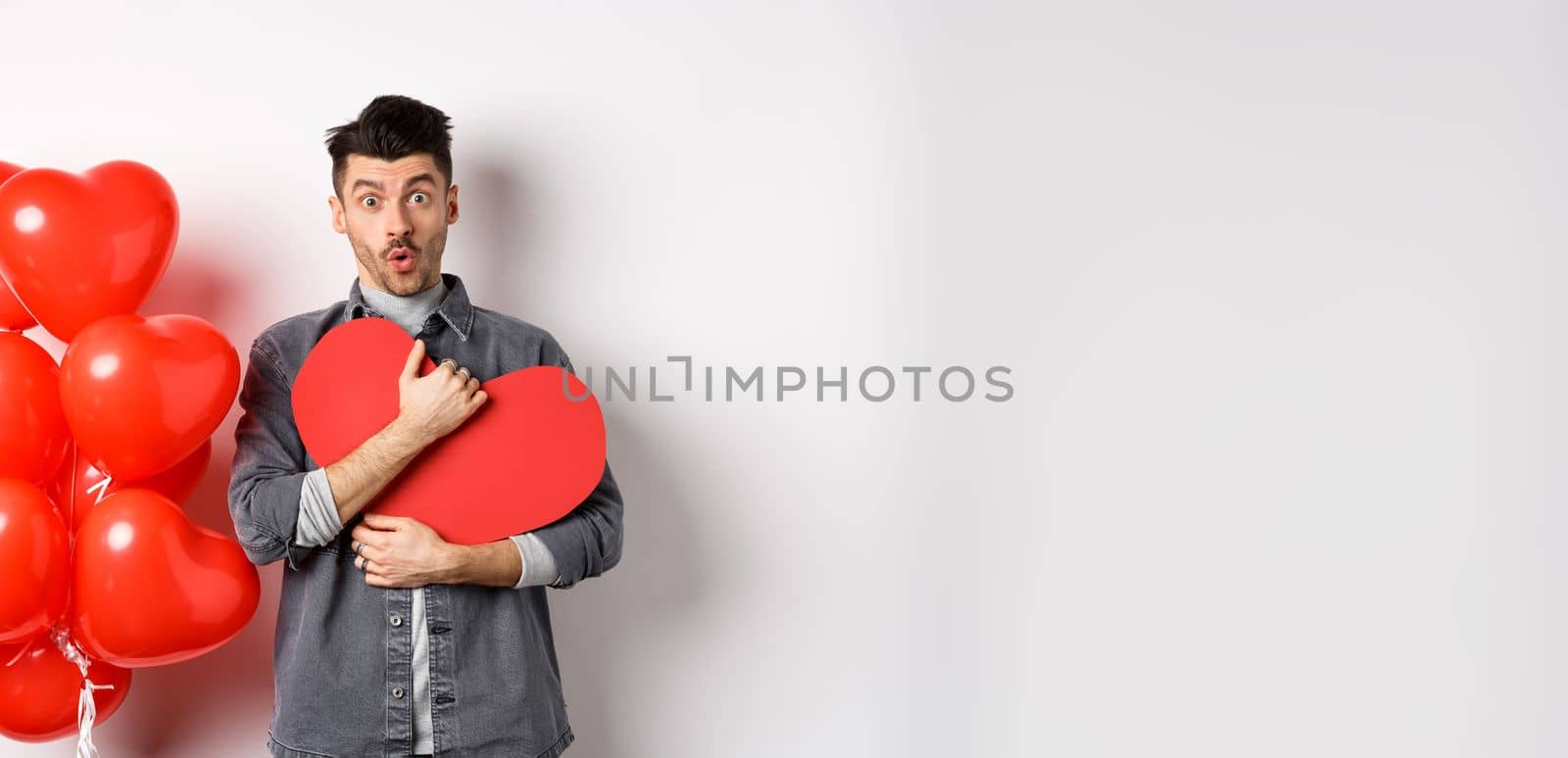 Surprised man holding Valentines heart card and saying wow, looking amazed at camera, receive secret confession on lovers day, standing near romantic balloons on white background by Benzoix