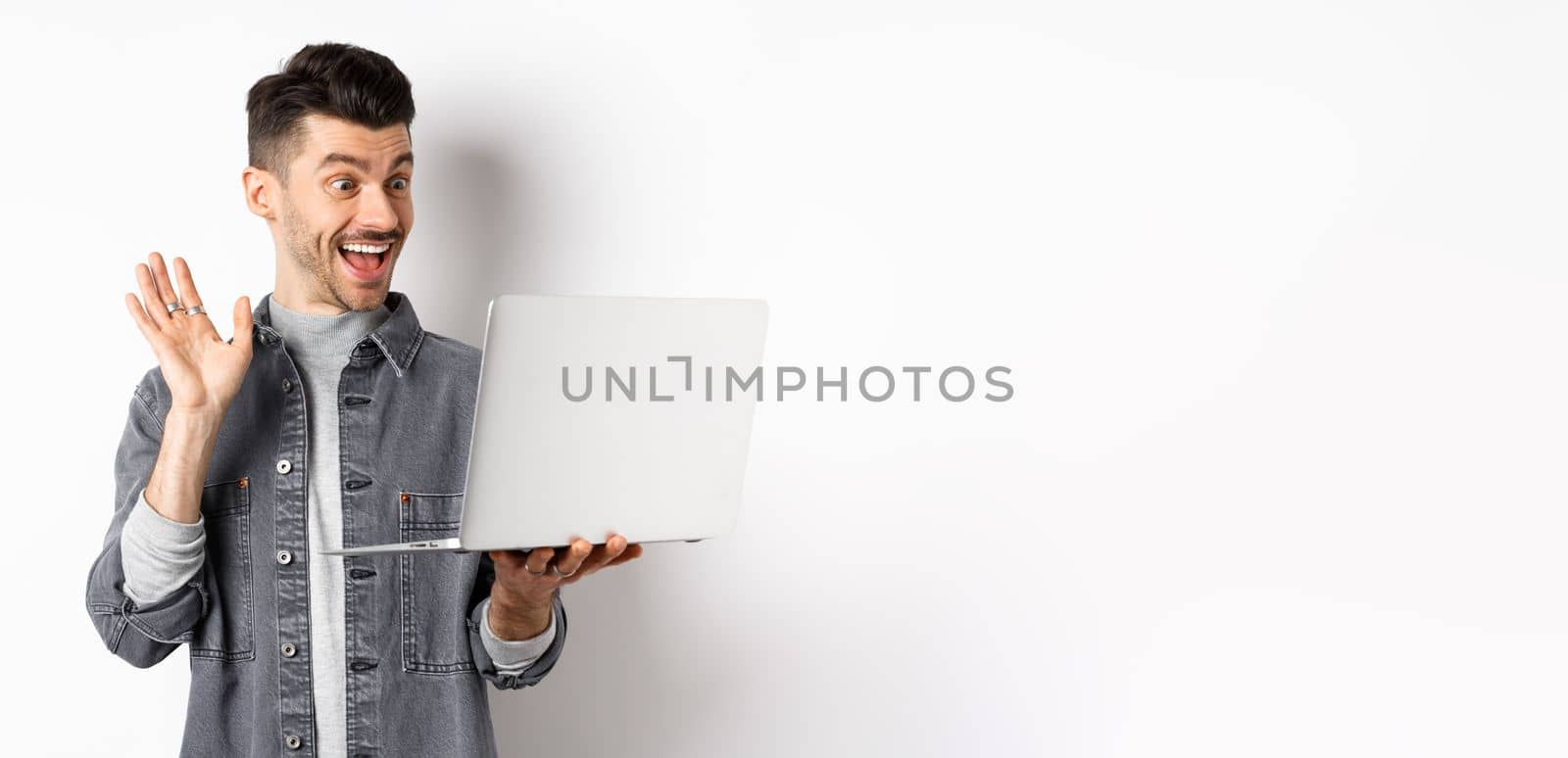 Excited man waiving hand at laptop, video chatting on computer and smiling friendly, standing on white background.