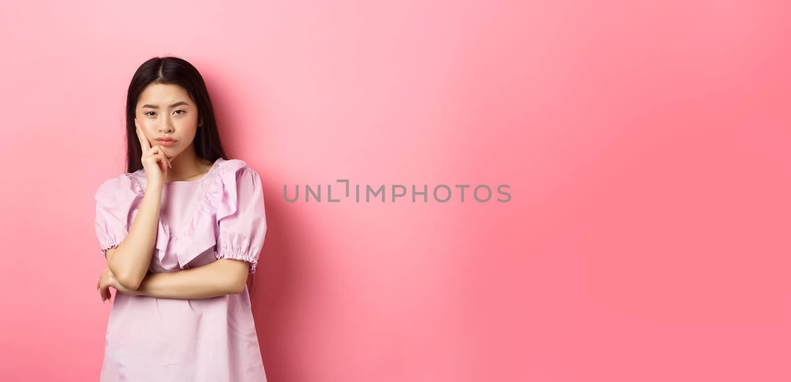 Bored asian teen girl look indifferent at camera, lean face on hand in skeptical pose, standing reluctant against pink background.