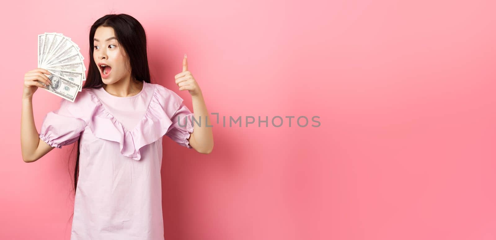 Excited asian girl looking at big money and make thumbs up, standing amazed with dollar bills against pink background.