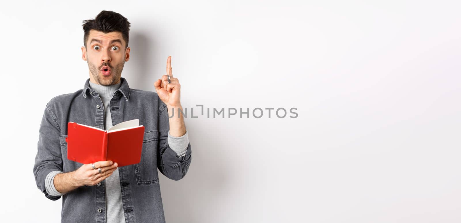Excited man pitch an idea while reading planner or diary, holding red journal and gasping amazed, raising finger in eureka sign, standing on white background.