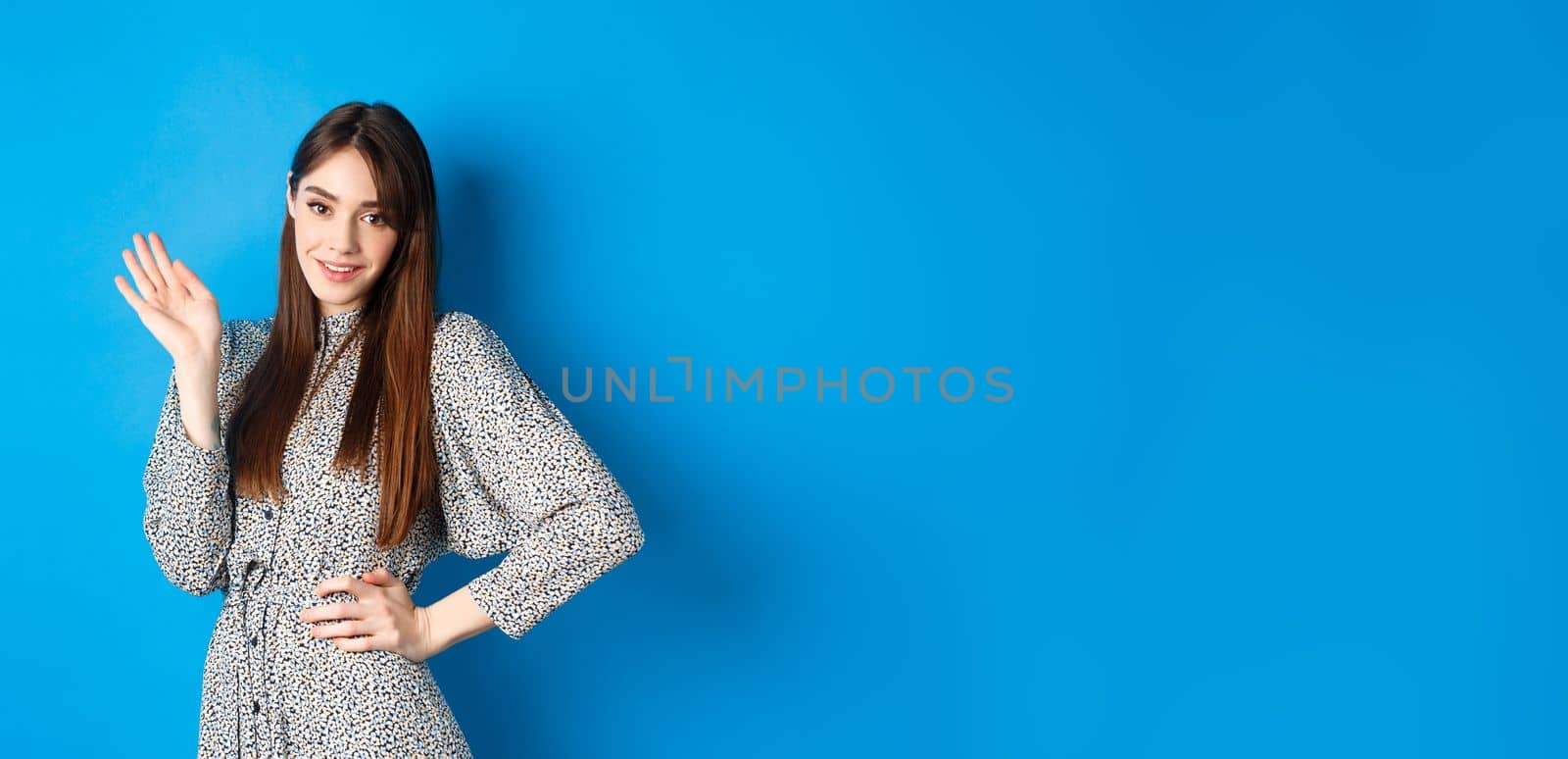 Beautiful lady in dress waving hand and say hello, greeting you with cute smile, standing on blue background by Benzoix