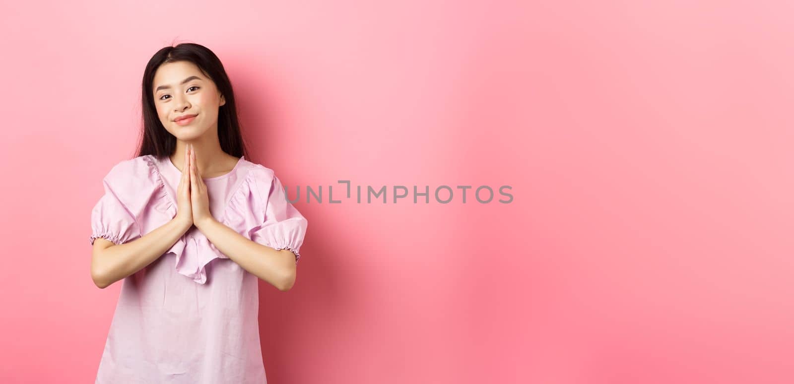 Cute asian girl say thank you, smiling and looking happy, showing namaste gesture in gratitude, standing in dress against pink background by Benzoix