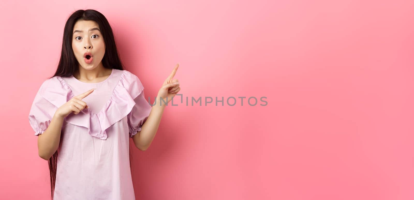 Excited asian woman look amazed, pointing right and gasping fascinated, checking out advertisement, standing on pink background by Benzoix