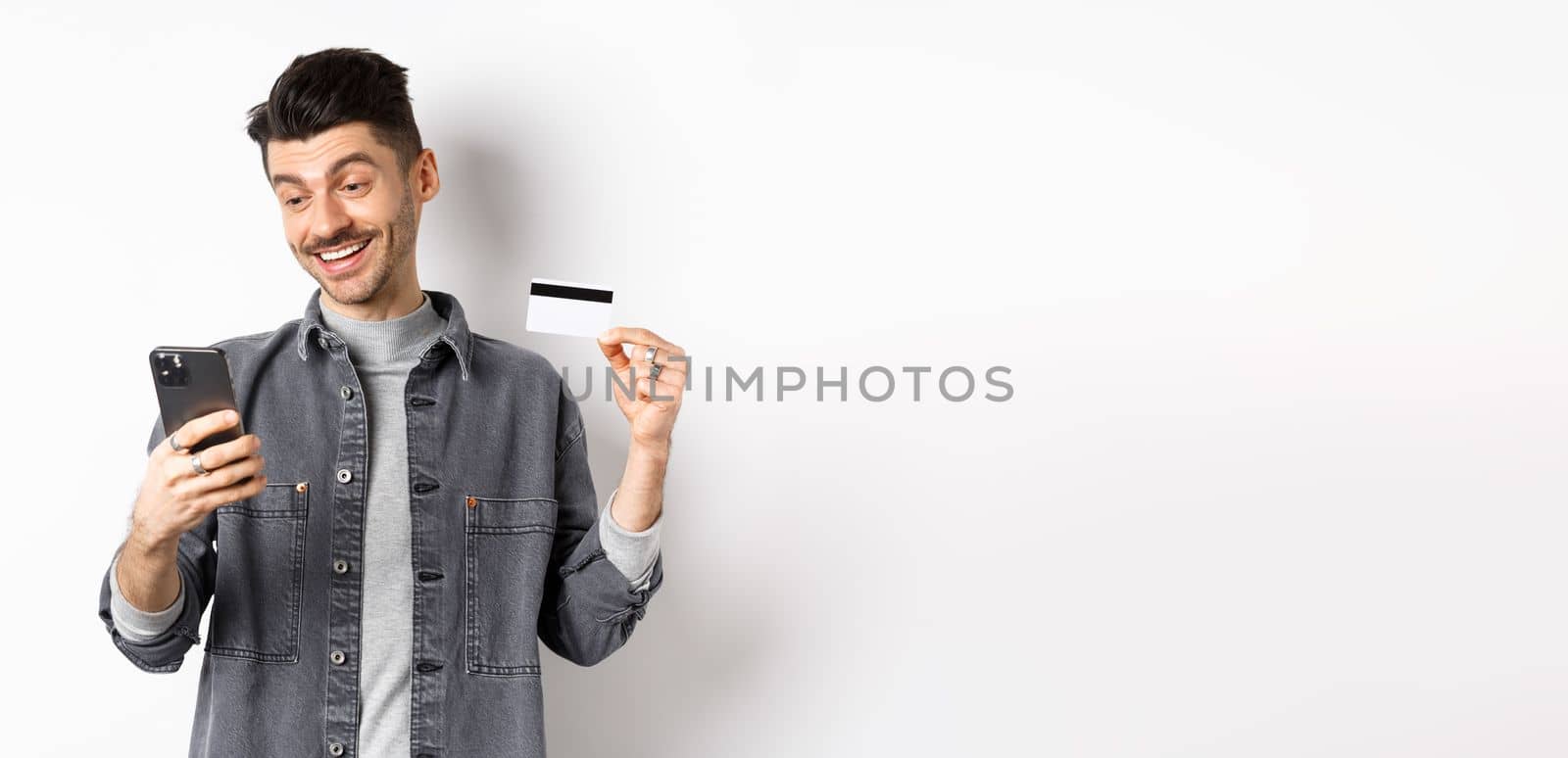 Online shopping concept. Handsome man buying in internet, showing plastic credit card and look at smartphone, standing against white background.